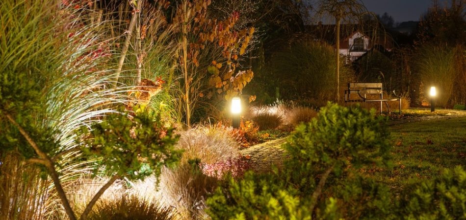 A garden path is illuminated by soft lights, surrounded by autumn foliage and a cozy bench under the evening sky, creating a serene atmosphere.