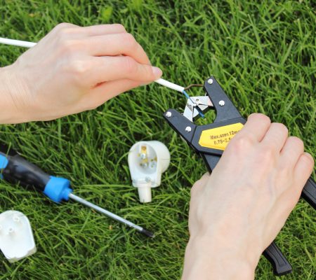 Hands stripping outdoor lighting wire with a tool on green grass beside a light fitting