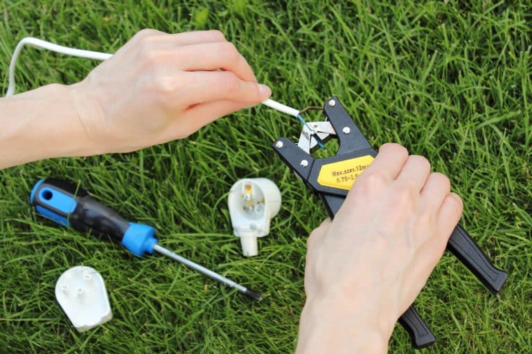 Hands stripping outdoor lighting wire with a tool on green grass beside a light fitting